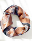 A rolled sunstone-colored silk cotton scarf with coral and navy botanical patterns cast in bright sunlight. Illustrated by Lisel Ashlock.
