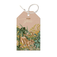 A beige gift tag with "FOR you" text and illustration of a brown monkey surrounded by green foliage and yellow flowers. Illustrated by Lisel Ashlock.