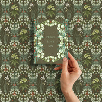 Hand holding a gift card with "Peace Love Joy" text over snowbell floral wrapping paper. Illustrated by Lisel Ashlock.