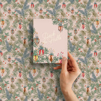 A hand holding a "Deck the Halls" greeting card against matching botanical holiday wrapping paper. Illustrated by Lisel Ashlock.