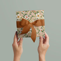 Hands holding floral gift wrap with brown ribbon bow against neutral background. Illustrated by Lisel Ashlock.