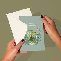 Hands holding an open Season's Greetings greeting card with blue background and floral illustration. Illustrated by Lisel Ashlock.