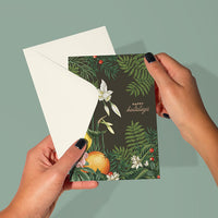 Hands holding open a dark green holiday greeting card with winter botanicals and citrus illustrations. Illustrated by Lisel Ashlock.