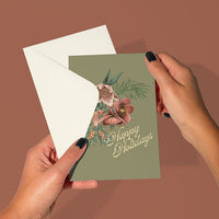 Hands holding open a mossy green greeting card with hellebore flowers and "Happy Holidays" text, illustrated by Lisel Ashlock.