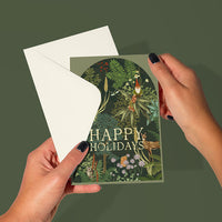 Hands holding an open "Happy Holidays" greeting card with woodland forest and botanical illustration by Lisel Ashlock.