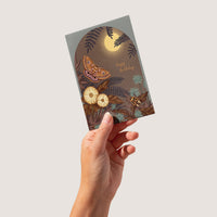 A hand holds an illustrated birthday greeting card featuring an autumn night scene with a moth, ferns, flowers, and moon. Illustrated by Lisel Ashlock.