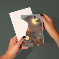 A hand holding an illustrated birthday greeting card featuring a moon, ferns, flowers, and an owl. Illustrated by Lisel Ashlock.