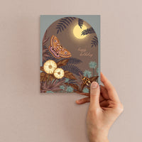 A hand holding a birthday greeting card featuring a nocturnal forest scene with a full moon, moth, flowers, ferns, and owl. Illustrated by Lisel Ashlock.
