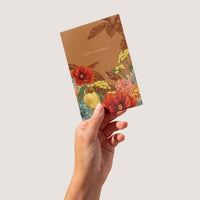 Hand holding a rust-colored birthday greeting card with illustrated red poppies, yellow flowers, and botanical elements. Illustrated by Lisel Ashlock.