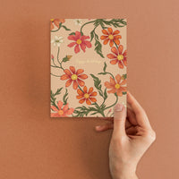 A hand holding a beige greeting card with red and orange cosmos flowers and green stems, "happy birthday" text centered. Illustrated by Lisel Ashlock.