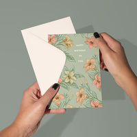 Hands holding open a floral birthday greeting card with pink and yellow flowers on sage green background. Illustrated by Lisel Ashlock.