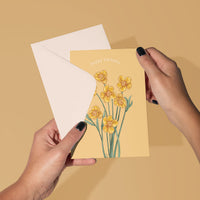 A person's hands holding an open yellow greeting card with yellow buttercup flowers and "happy birthday" text. Illustrated by Lisel Ashlock.