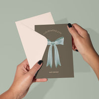Hands holding a "Happy Birthday" greeting card with "celebrating you" text and a blue bow illustration on taupe background. Illustrated by Lisel Ashlock.