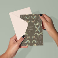 Hands holding open a Happy Birthday greeting card with dried flower design on taupe background. Illustrated by Lisel Ashlock.