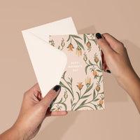 Hands holding a Mother's Day greeting card with floral design featuring yellow flowers and green leaves. Illustrated by Lisel Ashlock.