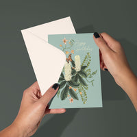 Person holding open a Happy Birthday greeting card with botanical illustrations of white flowers and a butterfly on sage green background. Illustrated by Lisel Ashlock.