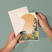 Person holding open a congratulations greeting card featuring an illustrated golden sunset with tropical foliage. Illustrated by Lisel Ashlock.