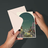 Hands holding open a "Thinking of You" greeting card featuring a teal night garden with illustrated flowers and butterflies. Illustrated by Lisel Ashlock.