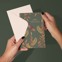 A hand holding an open "Giving Thanks" greeting card with botanical and moth illustrations on sage green background. Illustrated by Lisel Ashlock.