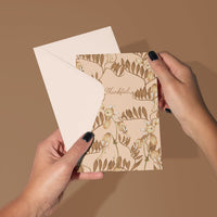 Hands holding a beige greeting card with "Thankful" text and botanical leaf design. Illustrated by Lisel Ashlock.