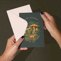 Hands holding open a dark green birthday greeting card featuring illustrated mushrooms, ferns, and a hare. Illustrated by Lisel Ashlock.