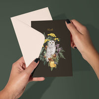 Hands holding a dark thank you greeting card featuring an illustrated snowy owl surrounded by yellow flowers and foliage. Illustrated by Lisel Ashlock.