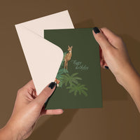 Hands holding open a green birthday greeting card featuring a deer on ferns with "happy birthday" text. Illustrated by Lisel Ashlock.