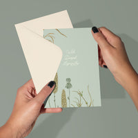 Hands holding an open sympathy greeting card with wildflowers and "With Deepest Sympathy" text. Illustrated by Lisel Ashlock.