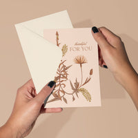 A hand holding a blush pink "Thankful For You" greeting card with botanical illustrations, envelope behind. Illustrated by Lisel Ashlock.
