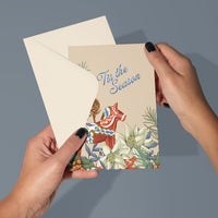 Hand holding a "Tis the Season" greeting card featuring a red Dala horse, pinecone, and holiday florals. Illustrated by Lisel Ashlock.