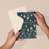 Hands holding a Happy Hanukkah greeting card with teal background and hanging flower illustrations. Illustrated by Lisel Ashlock.
