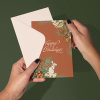 Hands holding open a "Happy Holidays" greeting card with rust-colored design featuring holly, pinecones, berries, and a white hare. Illustrated by Lisel Ashlock.