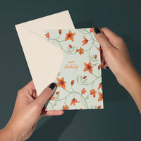 Hands holding a Happy Birthday greeting card with orange flowers and green vines on sea foam background. Illustrated by Lisel Ashlock.