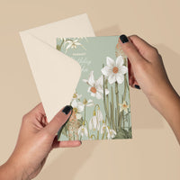 Hands holding an open birthday greeting card with white daffodils on sage green background. Illustrated by Lisel Ashlock.