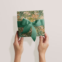 Two hands holding a square gift wrapped in pink paper with green monkeys and foliage, tied with a green ribbon bow. Illustrated by Lisel Ashlock.