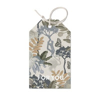 Gift tag with botanical illustration featuring plants, butterflies, and trees in blue, green, and tan on cream background with "FOR YOU" text. Illustrated by Lisel Ashlock.