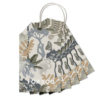 A fan of botanical gift tags with "FOR YOU" text, featuring illustrated plants, butterflies, and birds in muted tones. Illustrated by Lisel Ashlock.