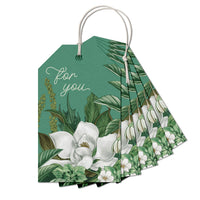 A fan-shaped stack of green gift tags with white magnolia flowers and "for you" text, tied with cream string. Illustrated by Lisel Ashlock.