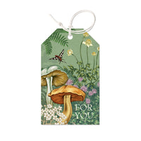 A woodland-themed gift tag featuring illustrated mushrooms, wildflowers, ferns, and a butterfly on green background with "FOR YOU" text. Illustrated by Lisel Ashlock.