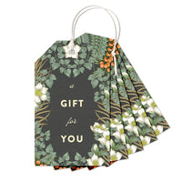 A fan-shaped gift tag with botanical illustrations of white flowers, green foliage, and orange berries on dark green background. Illustrated by Lisel Ashlock.