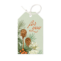 A mint green gift tag with "For You" in red script, decorated with pinecones, evergreen branches, berries, and white flowers. Illustrated by Lisel Ashlock.
