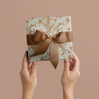 Two hands holding a wrapped gift with botanical floral pattern and brown ribbon bow against pink background, Illustrated by Lisel Ashlock.