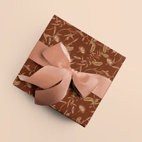 Rust-colored gift wrap featuring botanical lily tree pattern with pink ribbon bow on beige background. Illustrated by Lisel Ashlock.