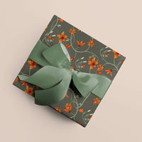 A wrapped gift box with charcoal gray wrapping paper featuring orange flowers and green vines, tied with a sage green ribbon bow. Illustrated by Lisel Ashlock.