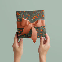 Two hands holding a wrapped gift box with dark green paper featuring orange floral patterns and a coral ribbon bow. Illustrated by Lisel Ashlock.