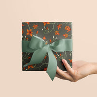 Hand holding dark gray gift wrap featuring orange flowers and green vines, tied with a sage green ribbon bow. Illustrated by Lisel Ashlock.
