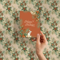 Hand holding terracotta greeting card with "Happy Holidays" text against festive holly and hare patterned background. Illustrated by Lisel Ashlock.