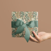 A hand holding a square gift wrapped in botanical patterned paper tied with a green ribbon bow. Illustrated by Lisel Ashlock.