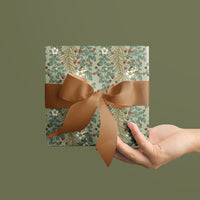 A hand holding wrapped gift with sage green floral and berry pattern tied with tan ribbon bow. Illustrated by Lisel Ashlock.