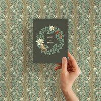 Hand holding a "Merry and Bright" holiday greeting card against coordinating sage gift wrap. Illustrated by Lisel Ashlock.